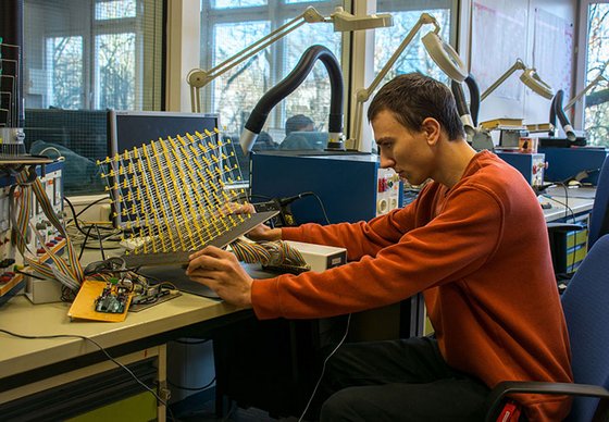 Ein junger Mann sitzt an einem Arbeitsplatz mit elektronischen Geräten und einem großen LED-Würfel.