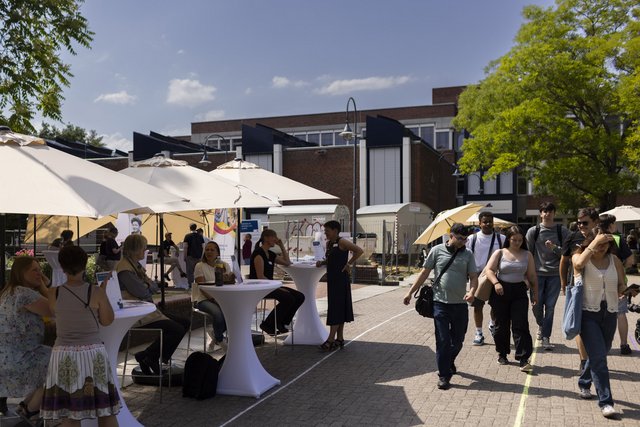 Personen sitzen an Infotischen unter Sonnenschirmen, junge Menschen laufen daran vorbei