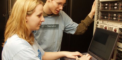 Zwei junge Menschen arbeiten in einem Serverraum an einem Laptop.