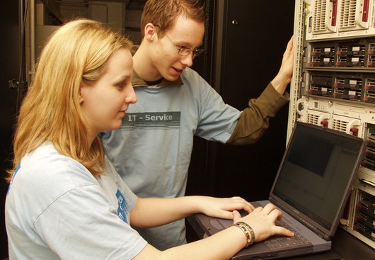 Zwei junge Menschen arbeiten in einem Serverraum an einem Laptop.