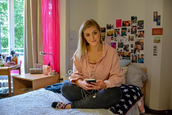 Eine junge Frau sitzt auf einem Bett und hört Musik über ihr Handy.