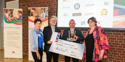 Drei Männer und eine Frau halten einen symbolischen Scheck, eine Urkunde und einen Pokal in die Kamera