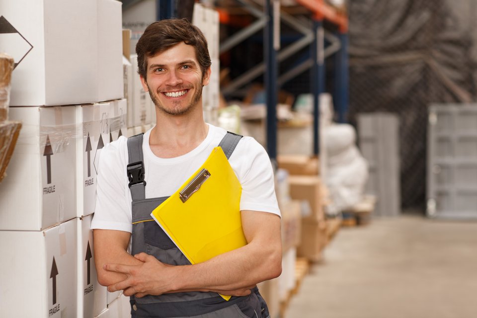Junger Mann mit Klemmbrett unter dem Arm lehnt seitlich an Boxen in einem Lagerraum.
