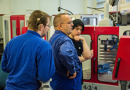 Zwei Auszubildende und ein Ausbilder in Arbeitskleidung in der Metallwerkstatt