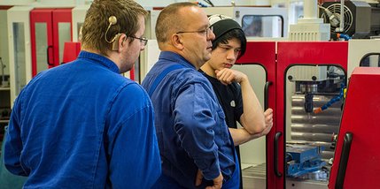 Zwei Auszubildende und ein Ausbilder in Arbeitskleidung in der Metallwerkstatt
