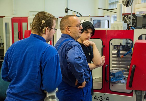 Drei Männer stehen in einer Werkhalle, einer davon trägt ein Cochlea Implantat.