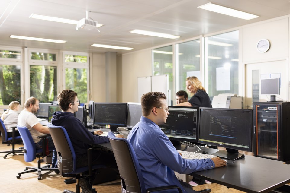 Vier Auszubildende arbeiten an ihren PCs, im Hintergrund erklärt eine Ausbilderin einem Auszubildenden etwas.