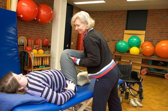 Eine Frau liegt auf einer Krankenliege. Ihr Bein wird von einer anderen Frau massiert und bewegt. Im Hintergrund liegen Medizinbälle.