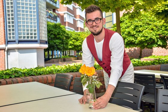 Ein junger Mann in Kellnerkleidung stellt eine Blumenvase auf einen Tisch.