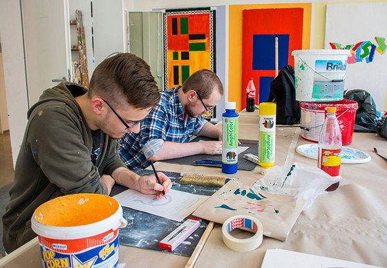 Zwei junge Männer sitzen an einem Tisch mit Farbeimern und Farbflaschen und zeichnen.