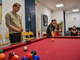 Zwei junge Männer spielen Billard, eine junge Frau schaut zu.