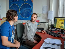 Ein junger Mann und ein älterer Mann sitzen in einem Büro vor dem PC und unterhalten sich.