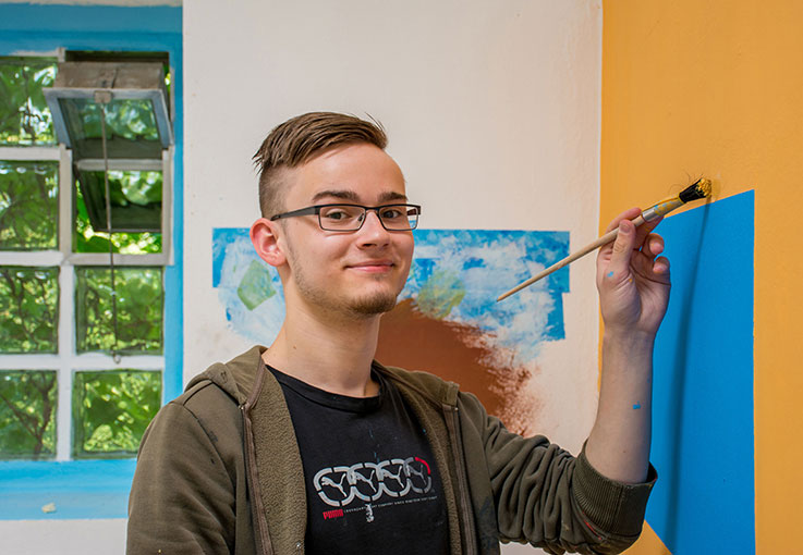 Eine Auszubildende für Maler und Lackierer hält einen Pinsel und malt eine Wand orange.