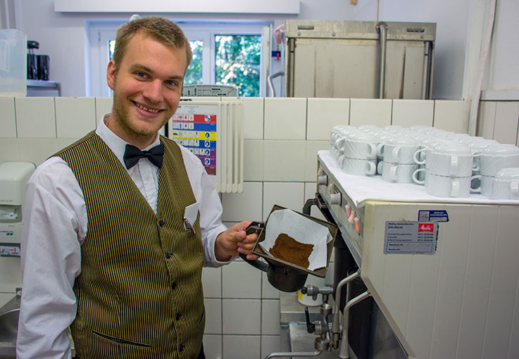 Ein Auszubildender im Service zeigt in der Küche frisch gemahlenen Kaffee und lächelt in die Kamera.