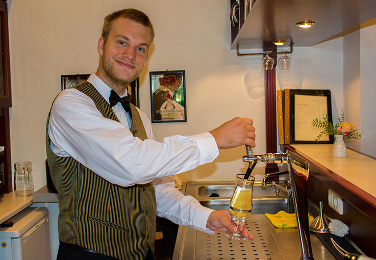 Ein Auszubildender mit Kellnerkleidung zapft ein Bier am Zapfhahn und lächelt in die Kamera.