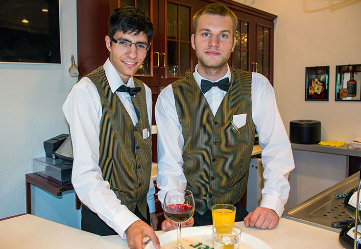 Zwei angehende Servicekräfte im Restaurant servieren Getränke und lächeln in die Kamera.