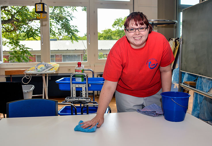 Eine Haushaltskraft wischt in einem Klassenraum einen Tisch ab und lächelt in die Kamera.