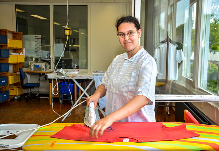 Eine Haushaltskraft bügelt ein T-Shirt und lächelt in die Kamera.