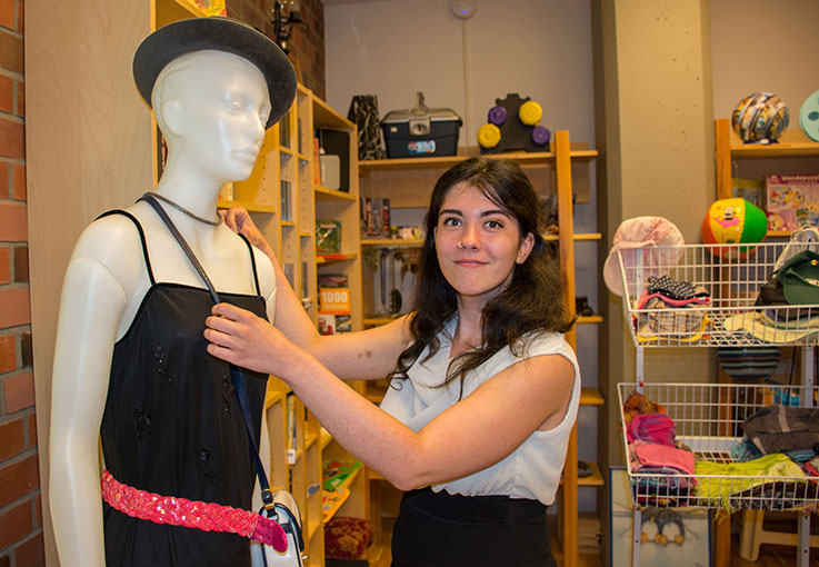 Eine Auszubildende dekoriert eine Schaufensterpuppe mit Accessoires und lächelt dabei in die Kamera.