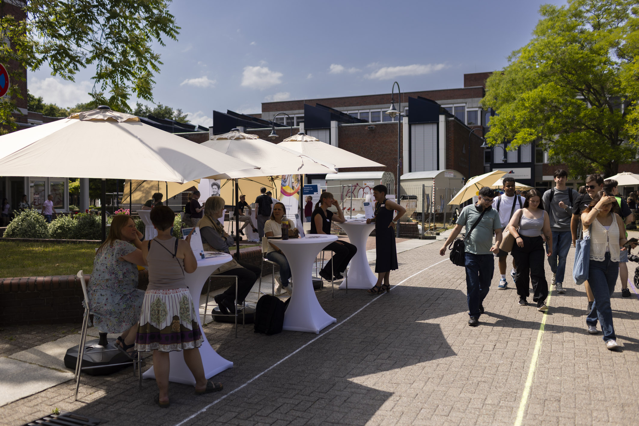 Personen sitzen an Infotischen unter Sonnenschirmen, junge Menschen laufen daran vorbei